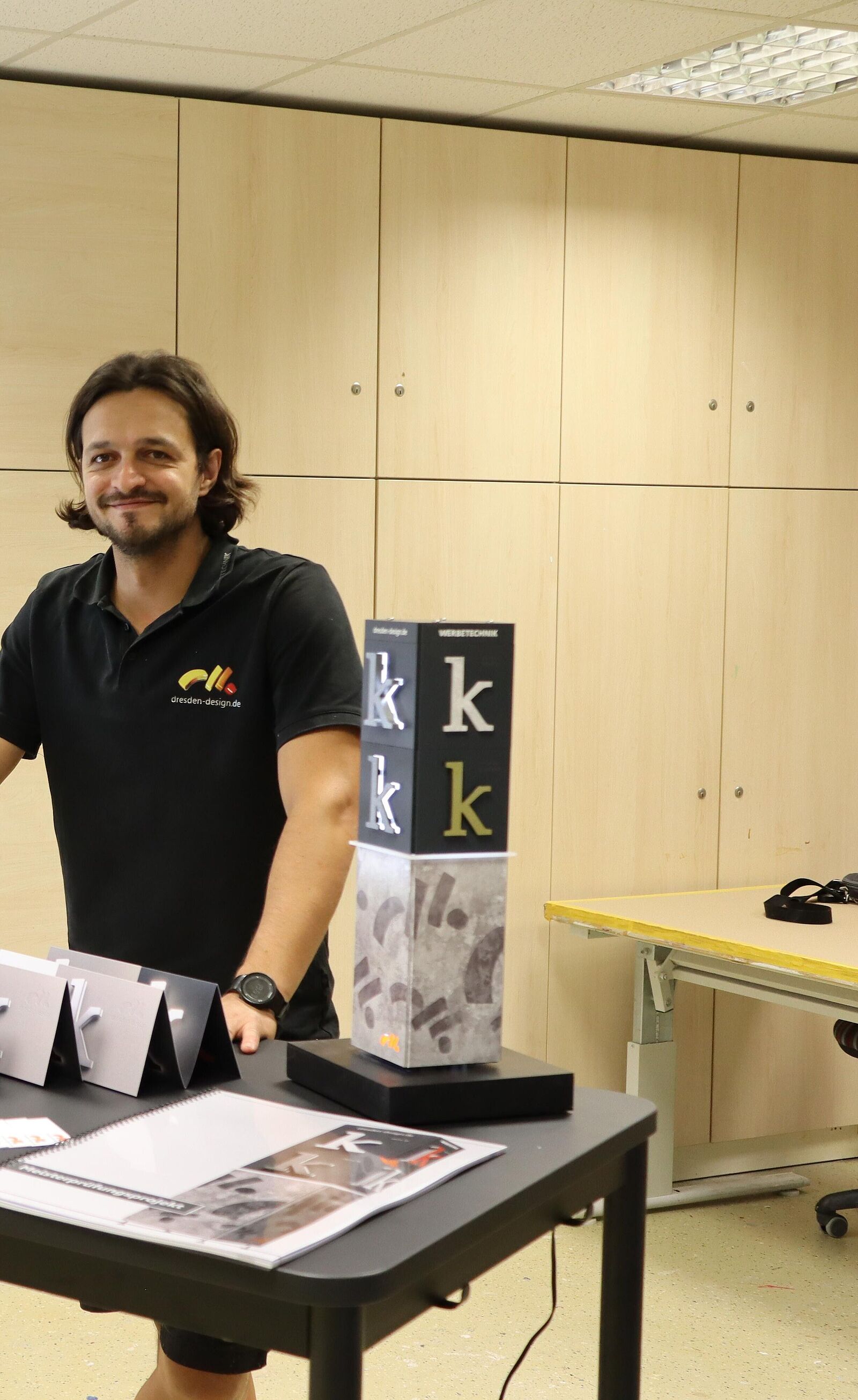 Ein Meister des Schilder- und Lichtreklamehersteller-Handwerks steht an einem Tisch vor seinen Meisterstücken: Reklamebuchstaben und verschiedenen Papier-Entwürfen, wie eine Reklamesäule.