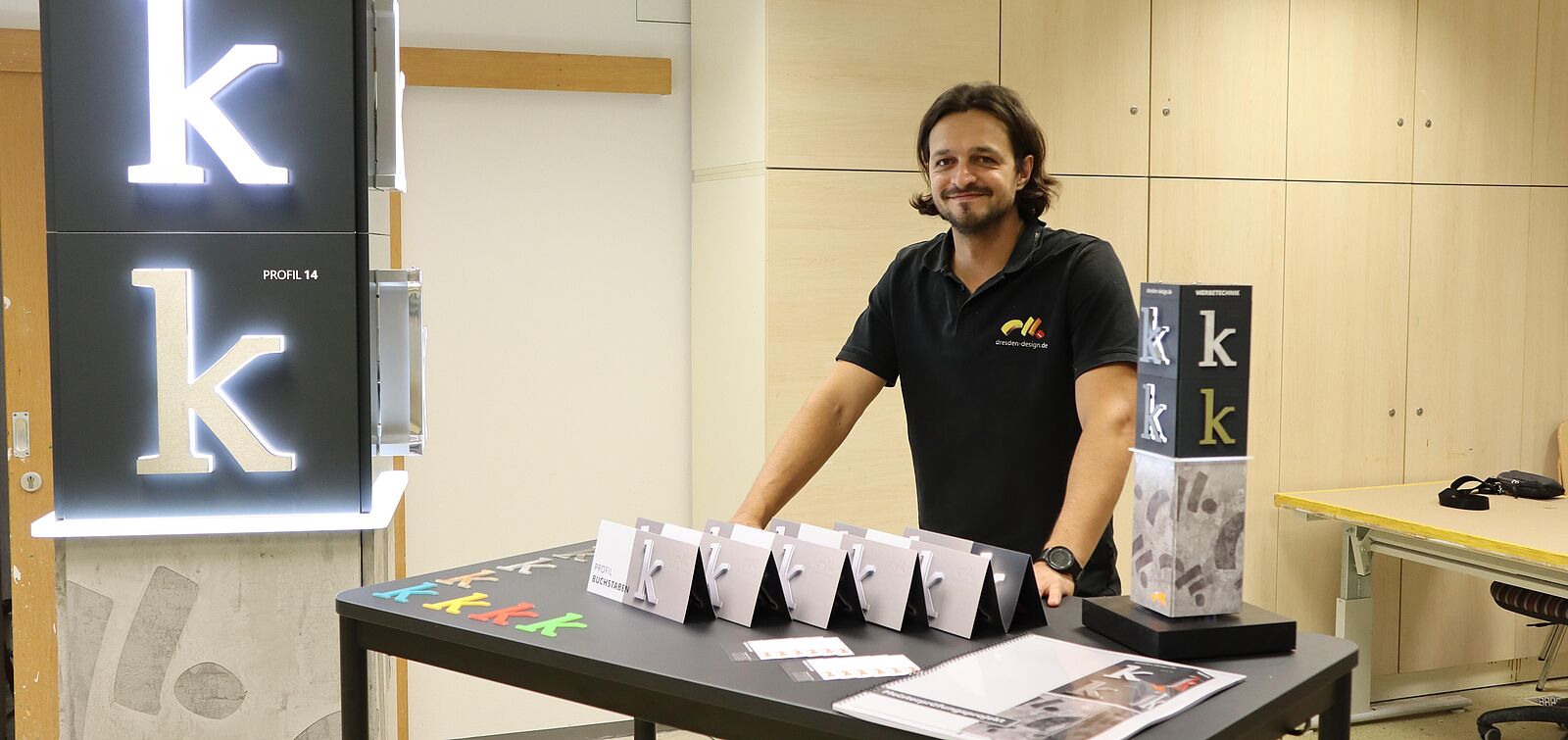 Ein Meister des Schilder- und Lichtreklamehersteller-Handwerks steht an einem Tisch vor seinen Meisterstücken: Reklamebuchstaben und verschiedenen Papier-Entwürfen, wie eine Reklamesäule.