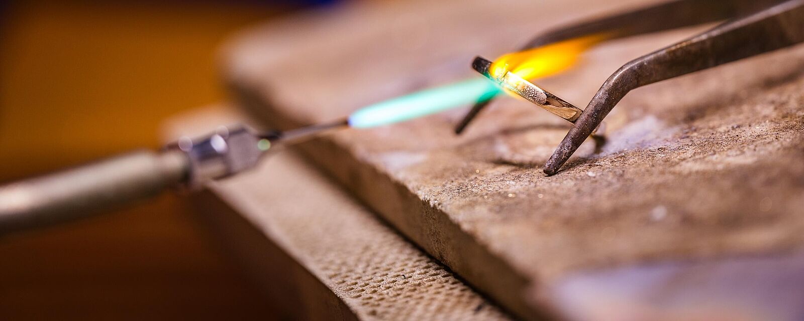 Nahaufnahme einer Goldbearbeitung in einer Werkstatt durch Pia Holz: Eine Zange hält einen Metallstab, der mit einer blauen Brennerflamme erhitzt wird. Der Hintergrund ist warm beleuchtet und unscharf.
