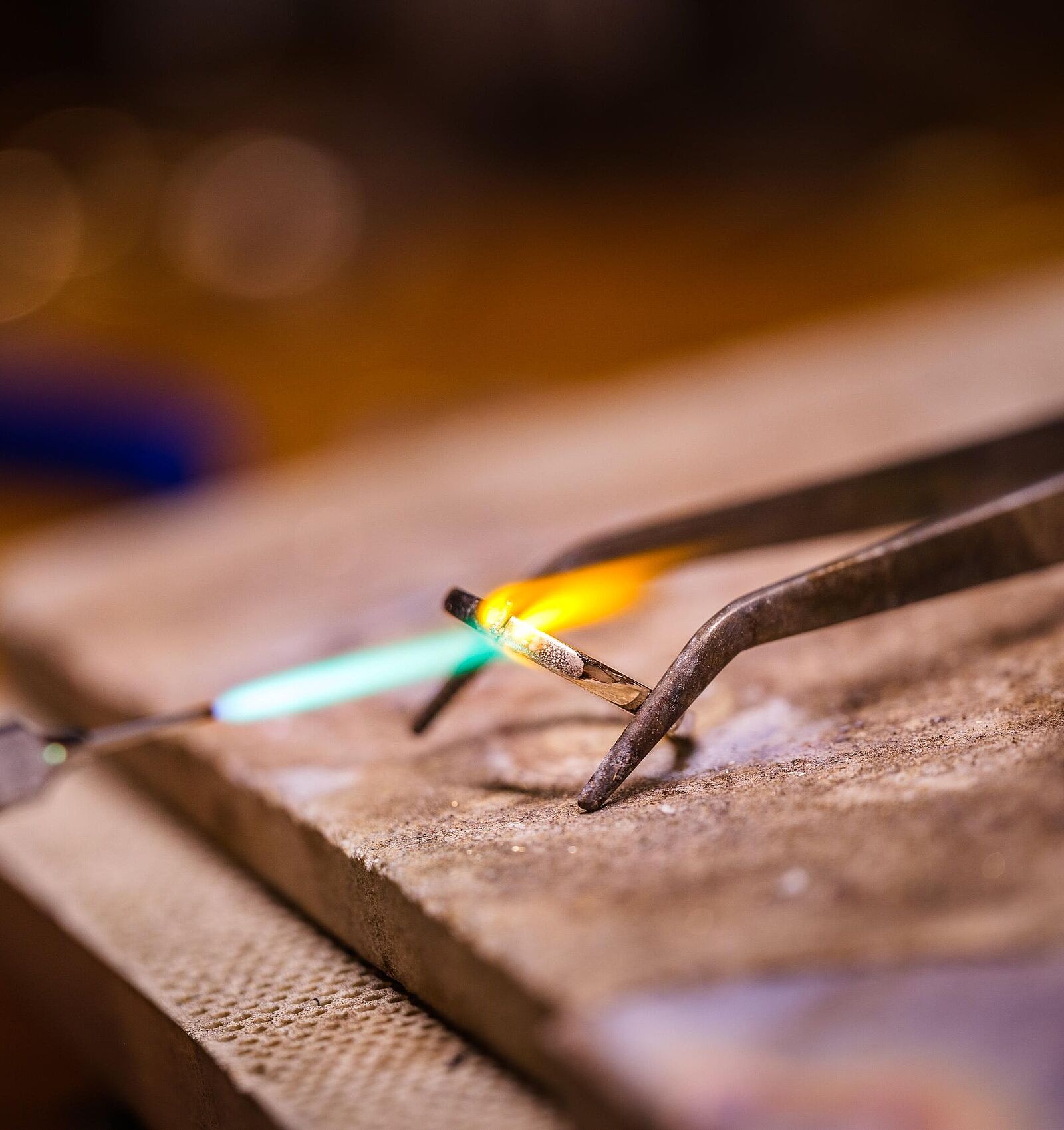 Nahaufnahme einer Goldbearbeitung in einer Werkstatt durch Pia Holz: Eine Zange hält einen Metallstab, der mit einer blauen Brennerflamme erhitzt wird. Der Hintergrund ist warm beleuchtet und unscharf.
