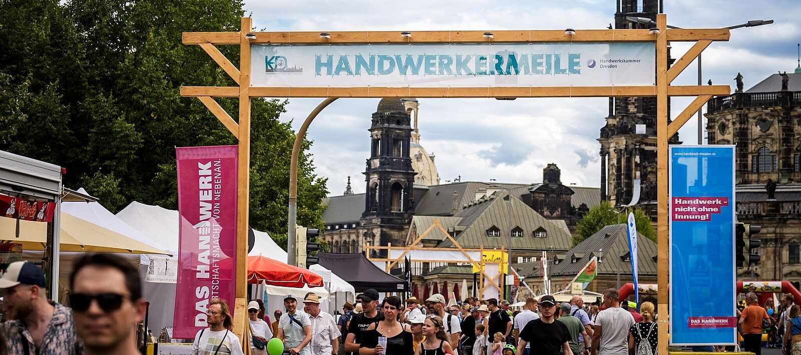 Menschen gehen durch ein hölzernes Tor mit dem Banner „Handwerkermeile\" der Handwerkskammer Dresden, dahinter Marktstände und die Dresdner Altstadtkulisse mit Kirchturm unter blauem Himmel.
