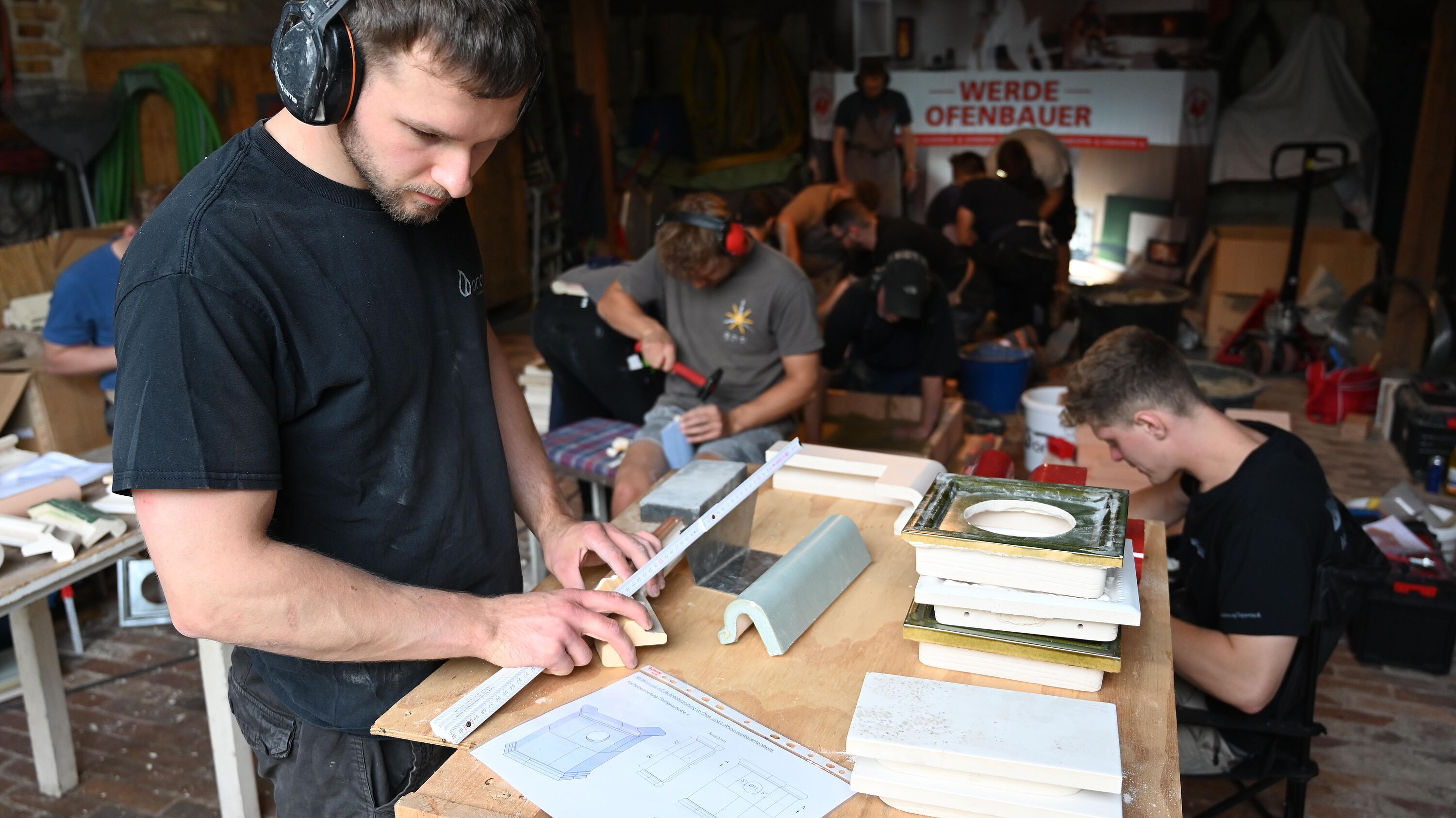 Der junge Handwerker, Erwin Otto, mit Gehörschutz misst in einer Werkstatt präzise ein geformtes Bauteil auf einer Holzarbeitsplatte. Auf dem Tisch liegen keramische oder steinerne Formteile, Werkzeuge und technische Zeichnungen. Im Hintergrund arbeiten weitere Auszubildende oder Handwerker an ähnlichen Werkstücken; ein Banner mit der Aufschrift „Werde Ofenbauer“ verweist auf Ausbildung und traditionelles Ofenbau-Handwerk. Das Bild zeigt konzentrierte Teamarbeit, handwerkliche Präzision, Ausbildung im Ofenbau sowie eine authentische Werkstattumgebung.
