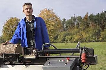 Eine Person steht auf einer Wiese mit einem großen Holzspalter, auf dem Holz liegt. Im Hintergrund ist ein Traktor zu sehen.