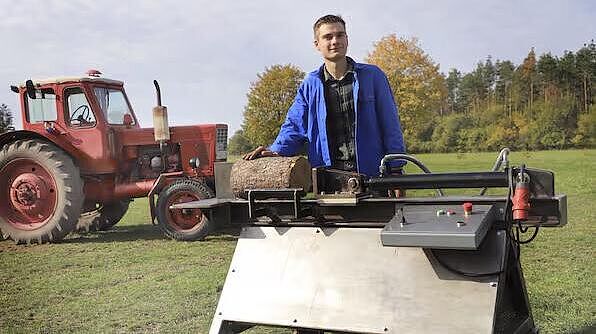 Eine Person steht auf einer Wiese mit einem großen Holzspalter, auf dem Holz liegt. Im Hintergrund ist ein Traktor zu sehen.