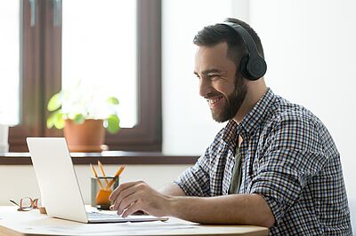 Eine Person sitzt mit Kopfhörern am Schreibtisch vor einem Laptop und verfolgt aktiv ein Webinar.