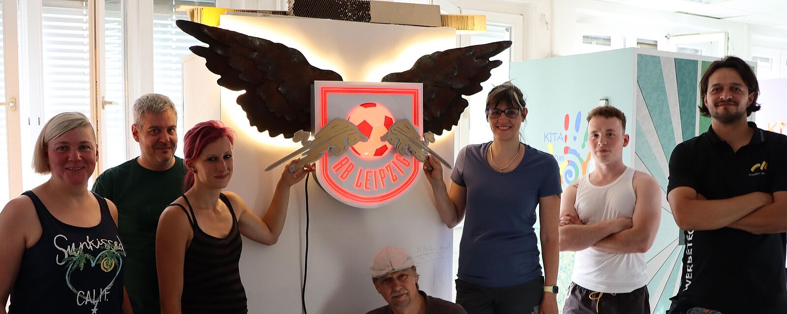 Gruppenaufnahme von Frauen und Männern einer Meisterklasse der Schilder- und Lichtreklamehersteller, vor ihrer Projektarbeit einem Logo des Fußballvereins RB Leipzig (als Kundenauftrag).