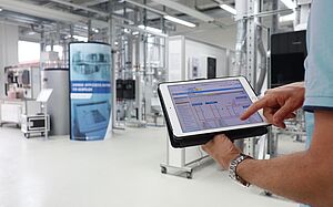 Blick in einen Werkraum des Energie-Effizienz-Zentrums, rechts im Vordergrund hält eine Person ein Tablet mit einer geöffneten App zum steuern von Geräten im Raum.