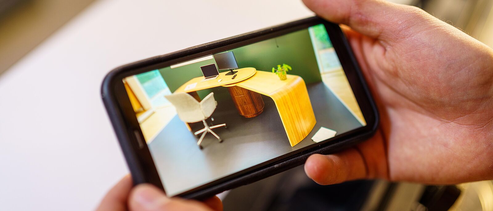 Makroaufnahme zweier Hände die ein Handy quer hält, im Display wird ein Gesellenstück ein Schreibtisch angezeigt.
