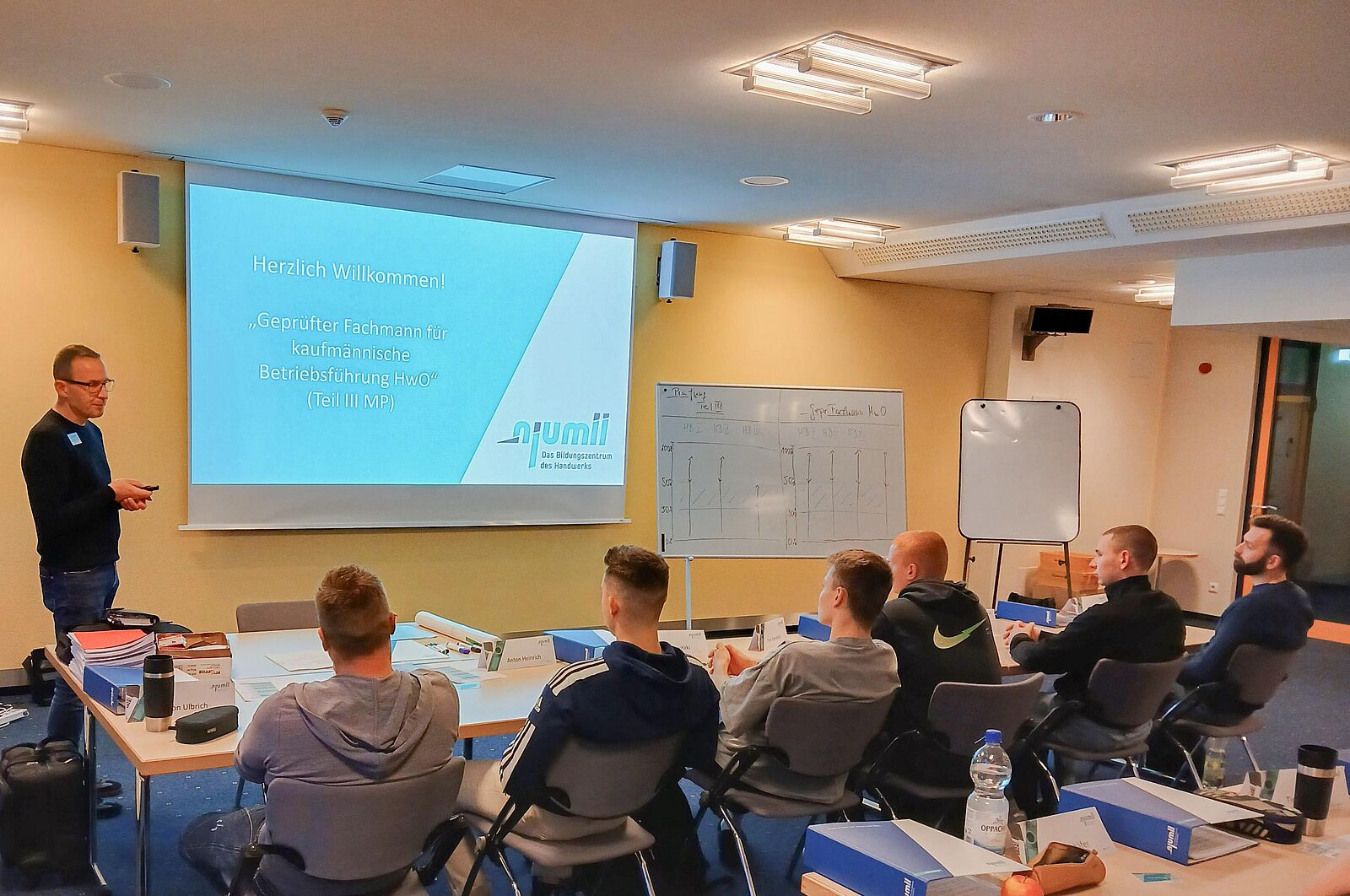 Blick in einen Unterrichtsraum auf die Präsentationswand. Ein Dozent hält einen Vortrag mit PowerPoint und stellt Fragen an die Zuhörer.