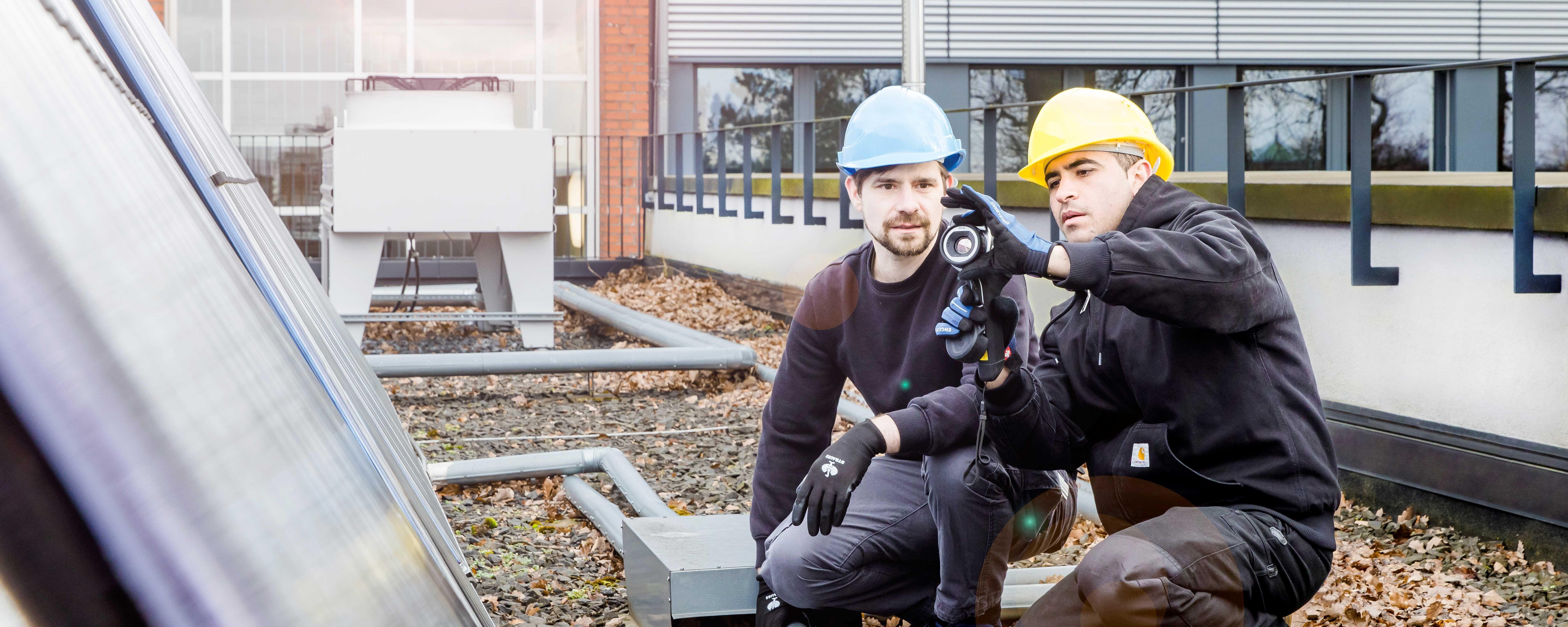 Zwei Personen mit Bauhelmen hocken auf einem mit grobem Kies bedeckten Dach. Eine Person hält ein Prüfgerät in der Hand und nimmt Einstellungen daran vor, die andere Person schaut zu und hilft.