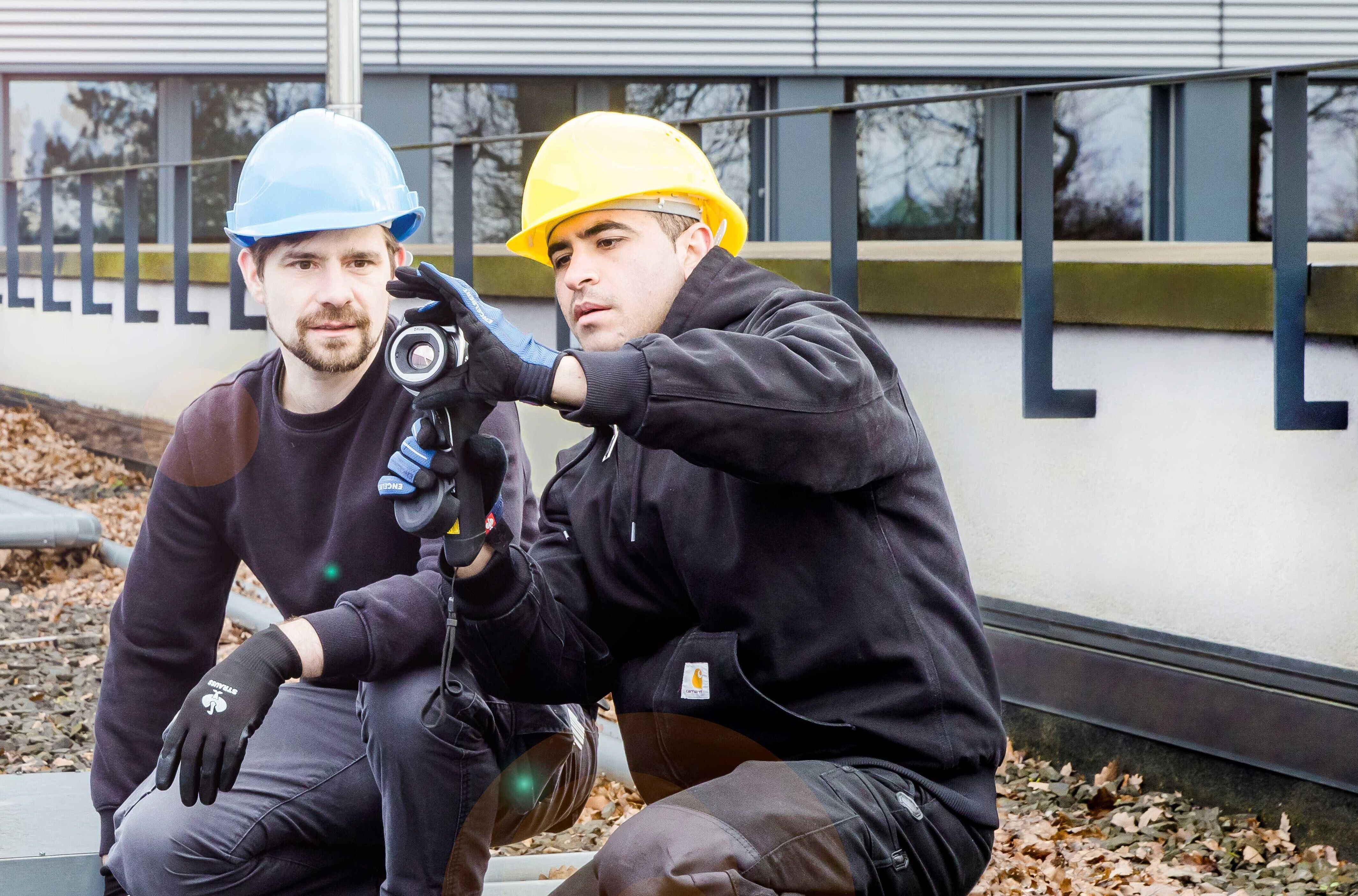 Zwei Personen mit Bauhelmen hocken auf einem mit grobem Kies bedeckten Dach. Eine Person hält ein Prüfgerät in der Hand und nimmt Einstellungen daran vor, die andere Person schaut zu und hilft.