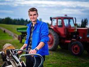 Eine Person steht mit ihrem Fahrrad auf einer Grün-/Ackerfläche, hinter ihr ein Traktor.