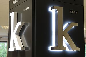 Makroaufnahme von einem Meisterstück eines Schilder- und Lichtreklameherstellers: Zwei weiße beleuchtete \"k\"-Reklamebuchstaben welche an einer eckigen Säule angebracht sind